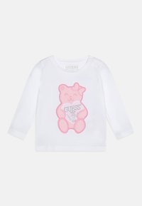 White long-sleeve shirt made of organic cotton, featuring a pink bear graphic with a heart and "GUESS?" text. Includes snap buttons at shoulder.