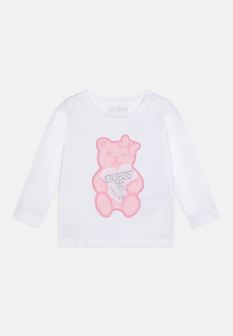 White long-sleeve shirt made of organic cotton, featuring a pink bear graphic with a heart and "GUESS?" text. Includes snap buttons at shoulder.