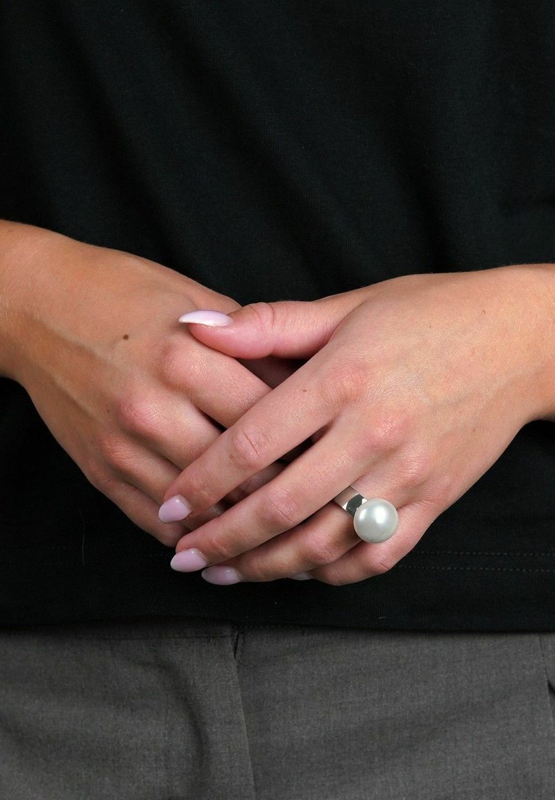 Silverring med en stor, rund, vit pärla på en slät band. Händer med ljusrosa naglar som är ihopknäppta, på en svart topp.