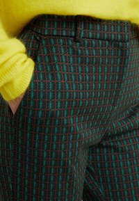 En person som bär gröna och bruna rutiga byxor med händerna i fickorna och en ljusgul fluffig tröja.