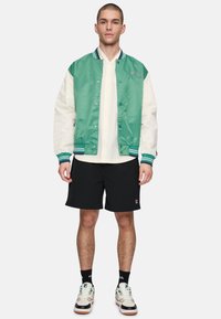 Giacca varsity in satin verde e bianco con colletto a righe, bottoni a pressione e logo ricamato, abbinata a pantaloni corti neri e sneaker.