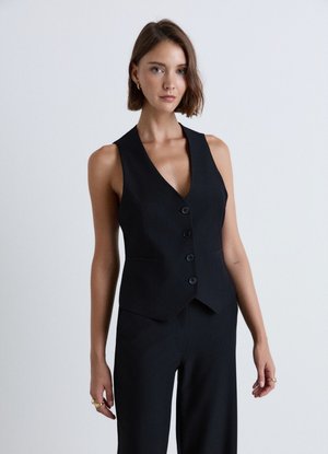 Donna con capelli castani corti che indossa un gilet nero senza maniche abbottonato e pantaloni neri su uno sfondo chiaro e uniforme.