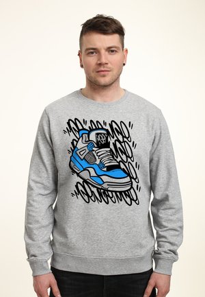 Młody mężczyzna w szarej bluzie z nadrukiem niebiesko-czarnego buta sportowego oraz abstrakcyjne czarne bazgroły w tle.