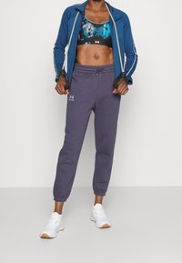 Blå lynlåsjakke, sort og blå sports-bh, grå sweats, hvide sneakers med orange detaljer, og fitnessudstyr i hånden.