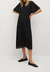 Schwarzes Midikleid mit einem gestuften, kurzärmligen Design. Hat Ausschnittdetails am Saum und wird mit schwarzen flachen Schuhen kombiniert.