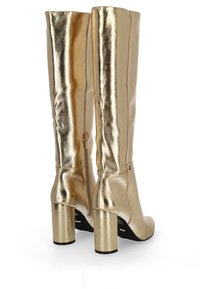 Wysokie metaliczne złote skórzane botki na słupku z bocznymi zameczkami, pokazane z tyłu na białym tle.