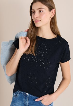 Jeune femme portant un haut noir à manches courtes perforé et un jean, tenant une veste en denim clair sur l'épaule, regardant vers la droite.