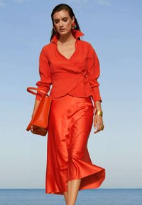 Rote Leinen-Wickelbluse mit langen Ärmeln, kombiniert mit einem roten Satin-Midirock. Die Accessoires umfassen eine geometrische rote Handtasche und auffällige Ohrringe.