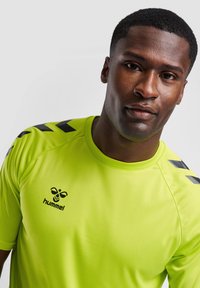 Camiseta deportiva de color verde neón, fabricada con tejido suave; presenta acentos en negro en los hombros y un logo en el lado izquierdo del pecho.