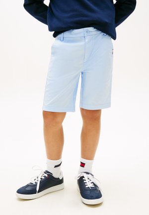 Bambino che indossa pantaloncini blu chiaro fino al ginocchio, sneakers blu navy con lacci bianchi, calzini bianchi e una felpa blu navy, in piedi su sfondo bianco.