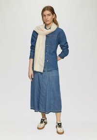 Jeansjack met een voorzak, gelaagd over een lange denimrok. Een gestructureerde roomkleurige sjaal drapeert om de nek. Lichtgekleurde sneakers maken de look compleet.