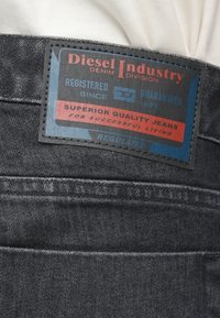 Tumši pelēni džinsa džinsi ar melnu un zilu etiķeti, kurā redzams teksts "Diesel Industry", sarkaniem akcentiem un reljefām detaļām.