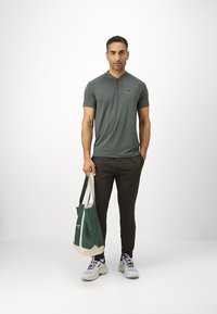 Grünes Poloshirt mit kurzen Ärmeln und Knopfkraagen, kombiniert mit dunkelgrünen Jogginghosen und weißen Turnschuhen; hält eine grüne Tragetasche mit cremefarbenen Akzenten.