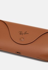 Écrin en cuir marron pour lunettes Ray-Ban avec fermeture à pression et un petit port de chargement sur fond blanc.