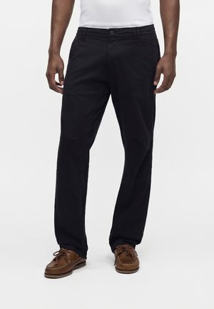 Homme portant un pantalon droit noir, des chaussures bateau en cuir marron et une chemise blanche, debout devant un fond blanc uni.