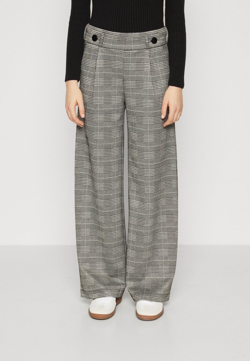 Femme portant un pantalon large taille haute à carreaux gris avec des boutons noirs, un haut noir côtelé à manches longues et des chaussures blanches, debout sur un sol gris.