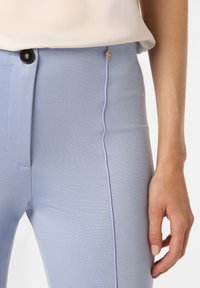 Hellblaue Hose aus strukturiertem Stoff, mit einem vorderen Knopfverschluss und einem dezenten goldenen Akzent in der Nähe des Bundes.