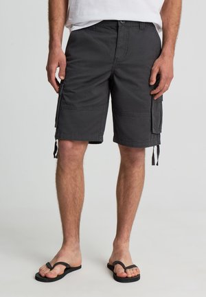 Uomo che indossa pantaloncini cargo neri, una maglietta bianca e infradito nere, in piedi davanti a uno sfondo chiaro e uniforme.