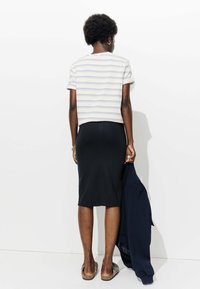 Femme aux cheveux bouclés courts se tient face à un mur blanc, portant un t-shirt à rayures, une jupe noire jusqu'aux genoux, des chaussures à enfiler à motifs, tenant une veste bleu marine.