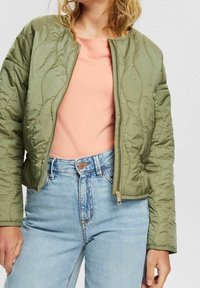 Olivgrüne, gesteppte Bomberjacke mit Reißverschluss, geripptem Kragen und Seitentaschen, getragen über einem hellrosa Hemd und hoch taillierten Jeans.