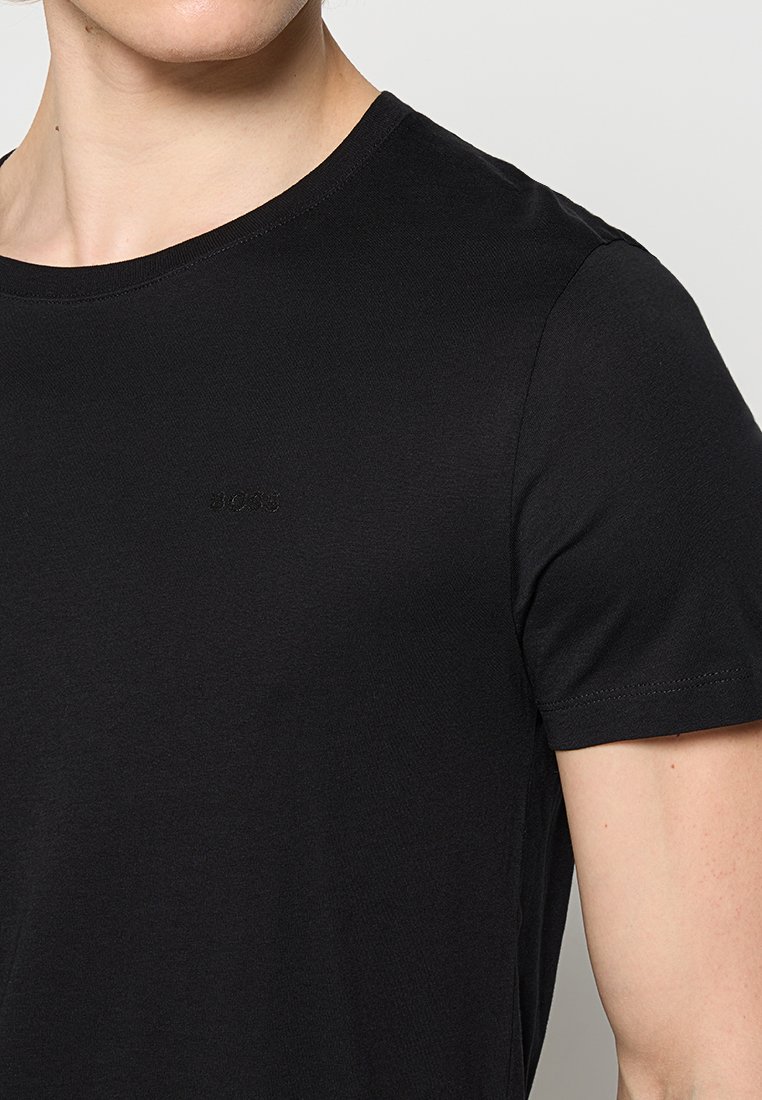 Nahaufnahme einer Person, die ein schlichtes schwarzes T-Shirt mit Rundhalsausschnitt trägt, auf dem sich dezent ein schwarzes besticktes Logo auf der Brust befindet.