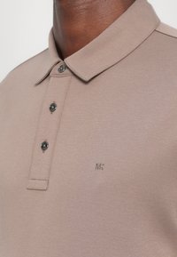 Béžové polo tričko z jemného materiálu s golierom, tromi tmavými gombíkmi a decentným vyšitým logom "MK" na ľavej strane.