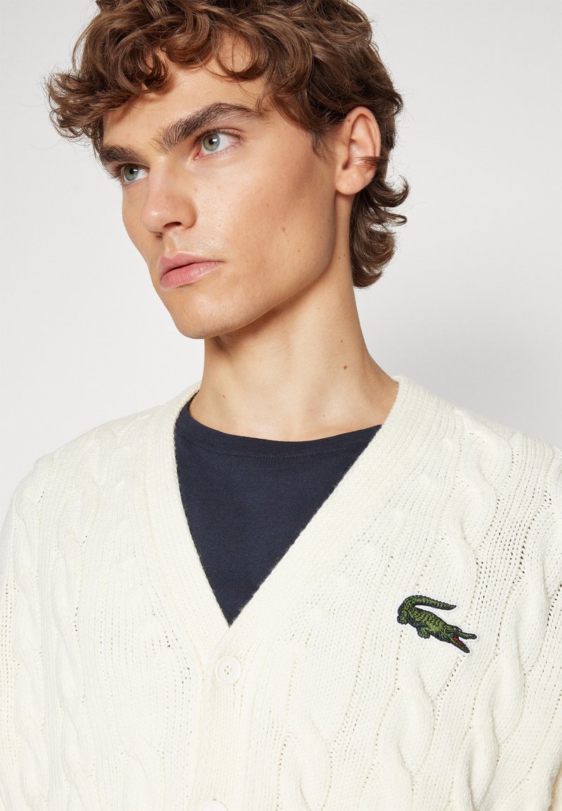 Cardigan crème en tricot torsadé avec un col en V, doté d'une fermeture à deux boutons et d'un logo crocodile vert brodé sur la poitrine gauche.