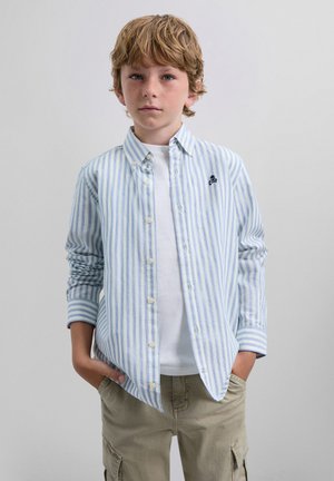 Camisa de botón con rayas verticales en azul y blanco, que presenta un pequeño diseño bordado en el pecho, combinada con pantalones cortos color caqui.