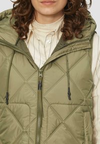 Olivgrüne gepolsterte Weste mit einem gesteppeten Rautenmuster, Reißverschluss, Kordelzugkapuze und zwei Fronttaschen. Weicher Stoff.