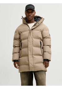 Chaqueta acolchada color arena con capucha, con un diseño acolchado, dos cremalleras laterales y acentos en negro contrastante. Usada sobre un suéter ligero.