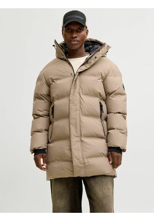 Tan pufferjack met een capuchon, voorzien van een doorgestikt ontwerp, twee zijkleppen en contrasterende zwarte accenten. Gedragen over een lichte trui.