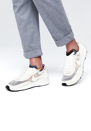 Pieds portant des baskets Nike blanches et grises avec des touches noires, assorties à un pantalon gris retroussé sur fond blanc.