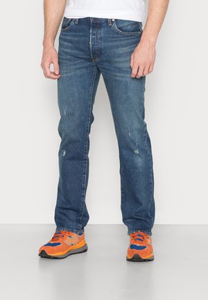 Homme portant un jean bleu délavé et des baskets orange vif et bleu, debout devant un fond clair uni.