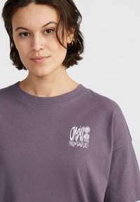 Paarse katoenen t-shirt met een ronde halslijn en korte mouwen. Het shirt heeft een witte grafische print op de borst met gestileerde tekst en bloemontwerpen.