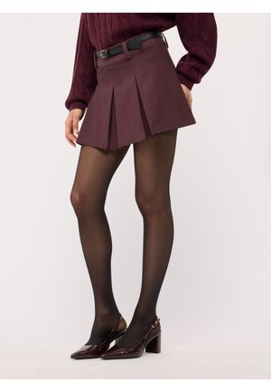 Donna che indossa una gonna plissettata viola scuro, cintura nera, collant neri trasparenti, maglione bordeaux e scarpe con tacchi color bordeaux, in piedi di lato.