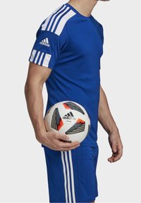 Blå idrottströja med vita accenter och tre ränder på ärmarna, kombinerad med blå shorts. En fotboll hålls i höger hand.