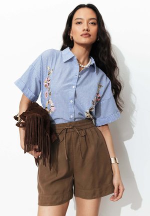 Femme portant une chemise à manches courtes brodée à rayures bleues, un short marron à cordon, tenant un sac marron à franges, avec des cheveux longs ondulés, un bracelet noir et un collier de perles.