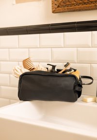 Zwarte leren toilettas met rits, gevuld met een houten kam, haarproducten en een spons, geplaatst op een witte wasbak.