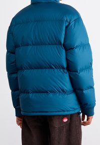 Tealfärgad vadderad jacka med hög krage, quiltad design, ribbade muddar och smal passform. Har en liten broderad logotyp på khakifärgade byxor.