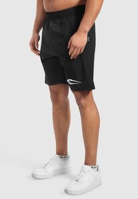 Pantalones cortos deportivos negros hechos de material ligero, con una cintura elástica y un logo blanco en el lado izquierdo. Combinados con zapatillas blancas.