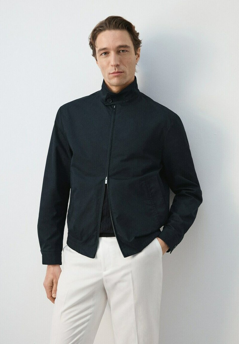 Homme portant une veste noire zippée avec un col boutonné et un pantalon blanc, debout devant un mur clair uni.