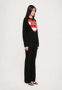 Kvinne står iført svarte bukser, svarte høyhælte sandaler og en svart genser med et stort rødt og hvitt hjerte med teksten "Moschino Jeans".