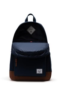 Námořnický modrý batoh s hnědým dnem, textilní materiál, kulatý tvar, přední kapsa se zipem a přední klopa zvýrazněná štítkem.