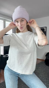 Vaaleanharmaa lyhythihainen t-paita, jossa on pyöreä pääntie; yhdistetty pastelliviolettiin ribbipipoon. Taustalla on moderni huone, jossa on pehmeitä tekstuureja.