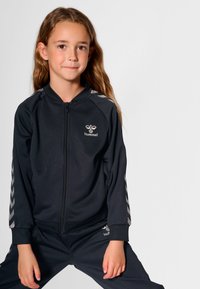 Czarny dres z kurtką zapinaną na zamek, ozdobioną srebrnymi akcentami w kształcie litery V na rękawach i logo na piersi, w komplecie z dopasowanymi spodniami.