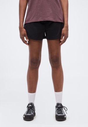 Person iført sorte shorts, en brun skjorte, hvide crew-sokker og grå og sorte vandresko stående mod en hvid baggrund.