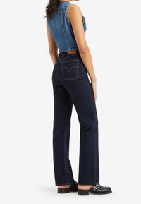 Femme portant un jean droit taille haute bleu foncé, un gilet en denim court sans manches et des mocassins noirs à boucle, debout sur fond blanc.