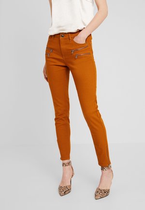 Mujer con pantalones ajustados color óxido con cuatro bolsillos delanteros con cremallera y tacones altos beige con estampado de serpiente, con la mano en el bolsillo.