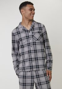 Jonge man die glimlacht, gekleed in een grijs en zwart geruit overhemd en bijpassende broek, staande met één hand in zijn zak tegen een effen achtergrond.