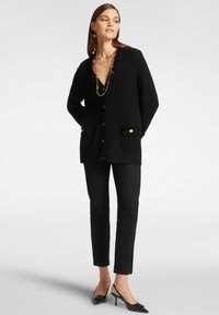 Cardigan nero in maglia con maniche lunghe, chiusura a bottoni, due tasche frontali e dettagli testurizzati. Abbinato a pantaloni neri aderenti e scarpe con tacco.